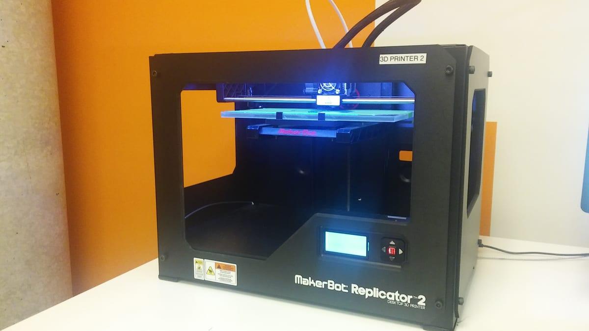 An enclosed black desktop 3D printer on a white bench against an orange wall, with a sticker on the top corner reading "3D PRINTER 2"