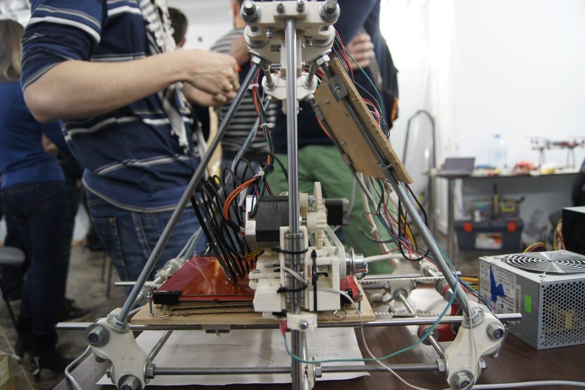 Several people gathered around a part-assembled open-frame 3D printer on a workshop table, one of them tightening a threaded rod, with a toolbox and a spare power supply beside it