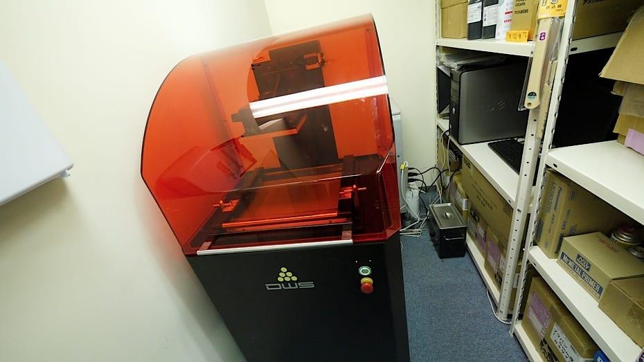 A large orange-hooded SLA resin 3D printer standing in a workshop beside shelves of boxed supplies