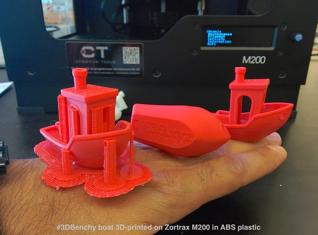 Three small red 3D printed boat test models in ABS held on an open hand in front of an enclosed desktop printer, the front one still attached to its support raft