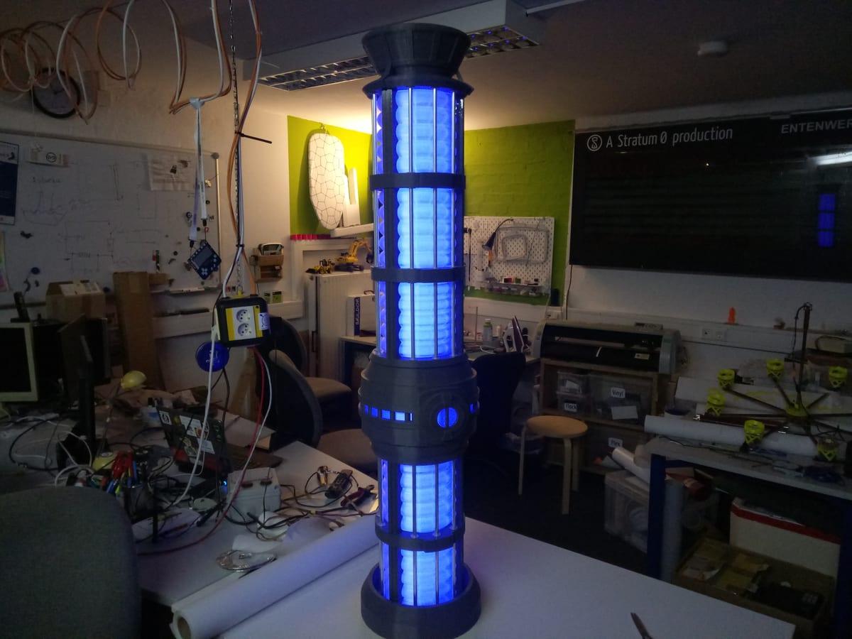A tall 3D printed column lamp in a hackerspace, built from stacked black printed rings and translucent panels lit blue from inside